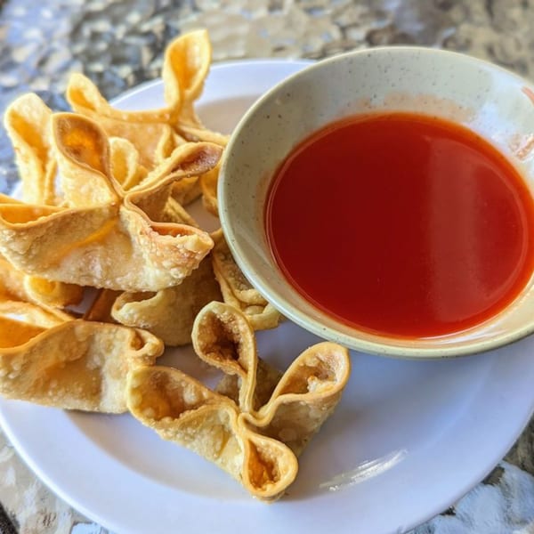 Crispy golden crab rangoons arranged on a white plate next to a bowl of red dipping sauce.